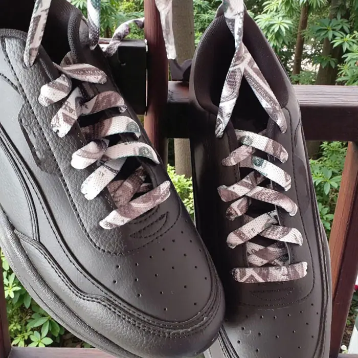 Czarne sneakersy wiszące na drewnianej balustradzie, ze sznurowadłami ARASHI US Dollar w biało-brązowym wzorze, w otoczeniu zieleni.